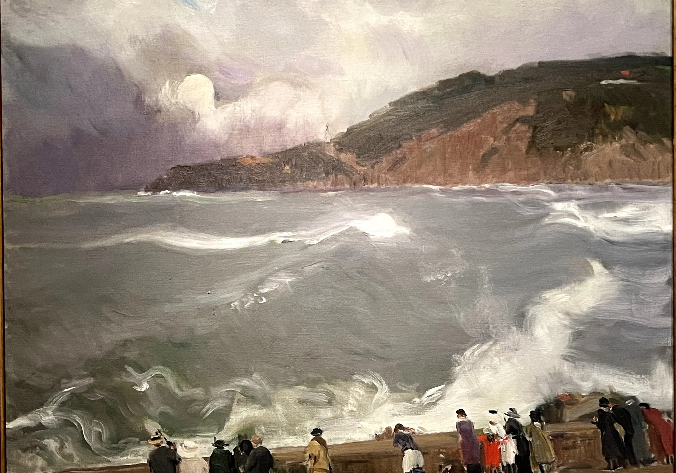A crowd in coats and hats gathers on a breakwater to watch a grey, storm-driven sea break in white spray beneath a headland — a dramatic late coastal scene under a cool northern sky.