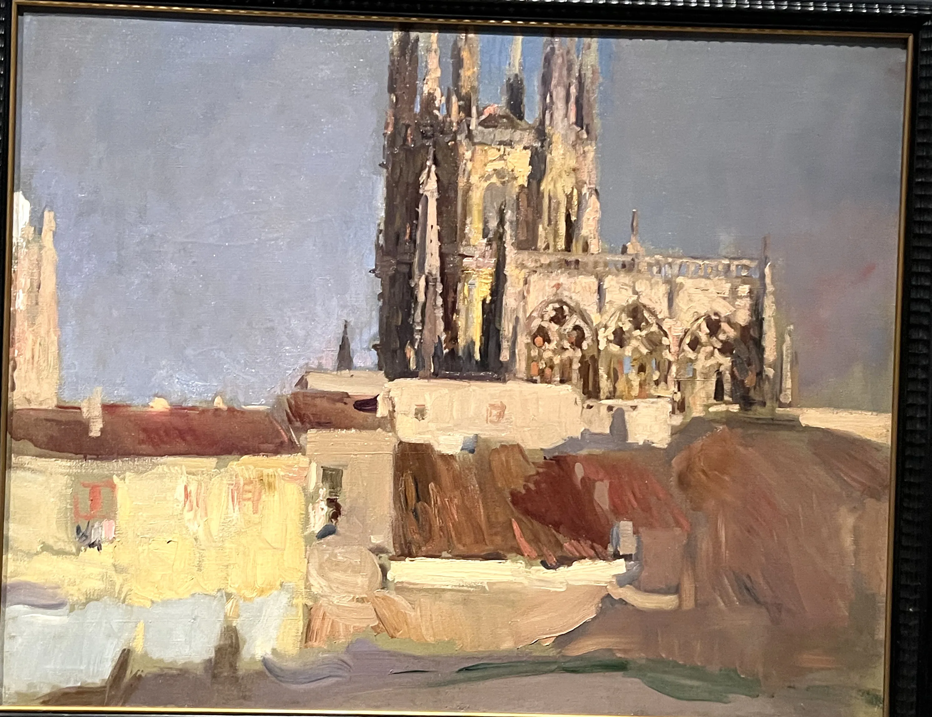 The pale Gothic spires and pinnacled facade of Burgos Cathedral catch the sun above a huddle of ochre and terracotta rooftops under a soft grey-blue sky.