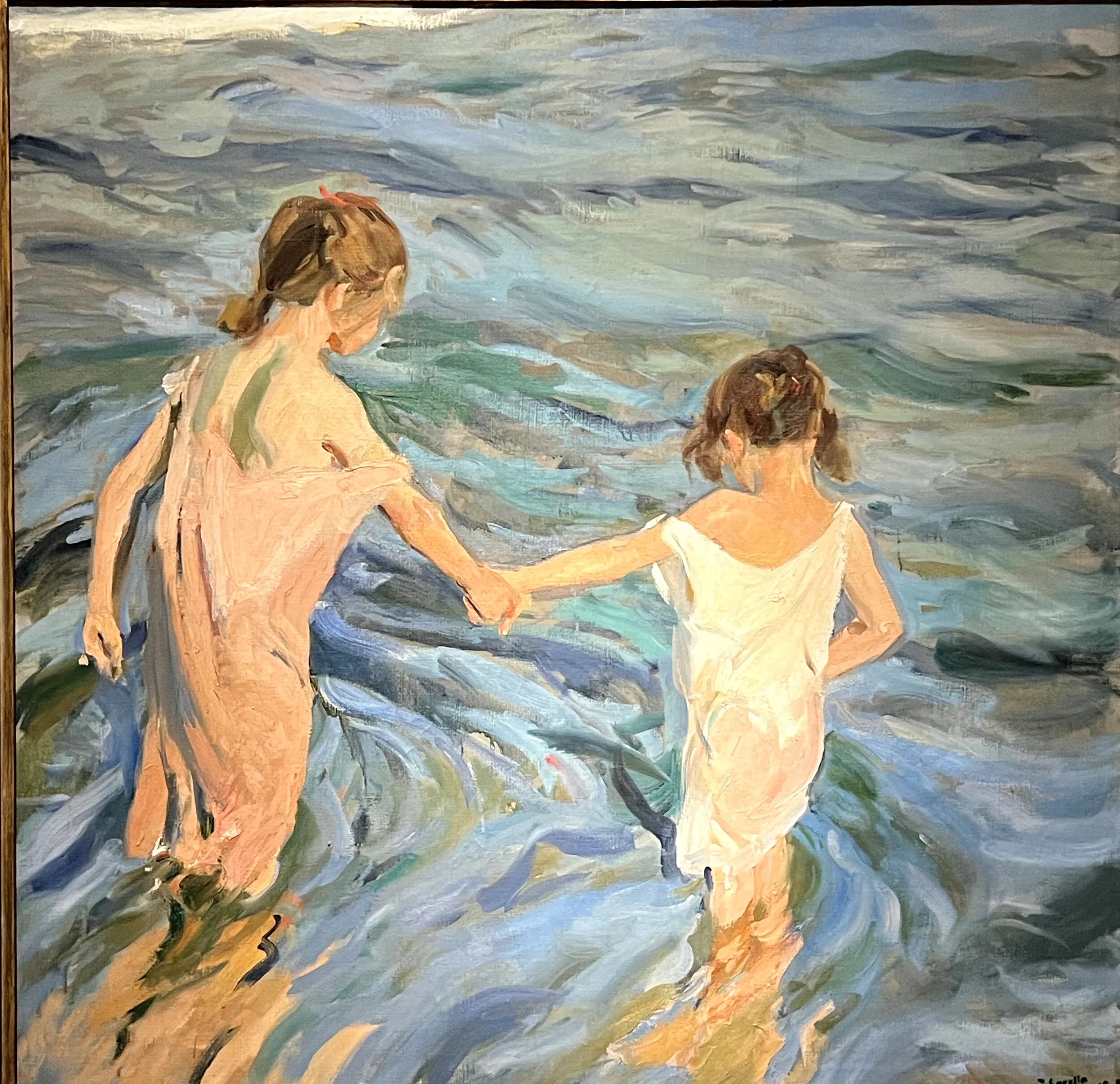 Two girls in white shifts, seen from behind and holding hands, wade into shallow surf whose swirling blues, greens and violets shimmer with reflected light.