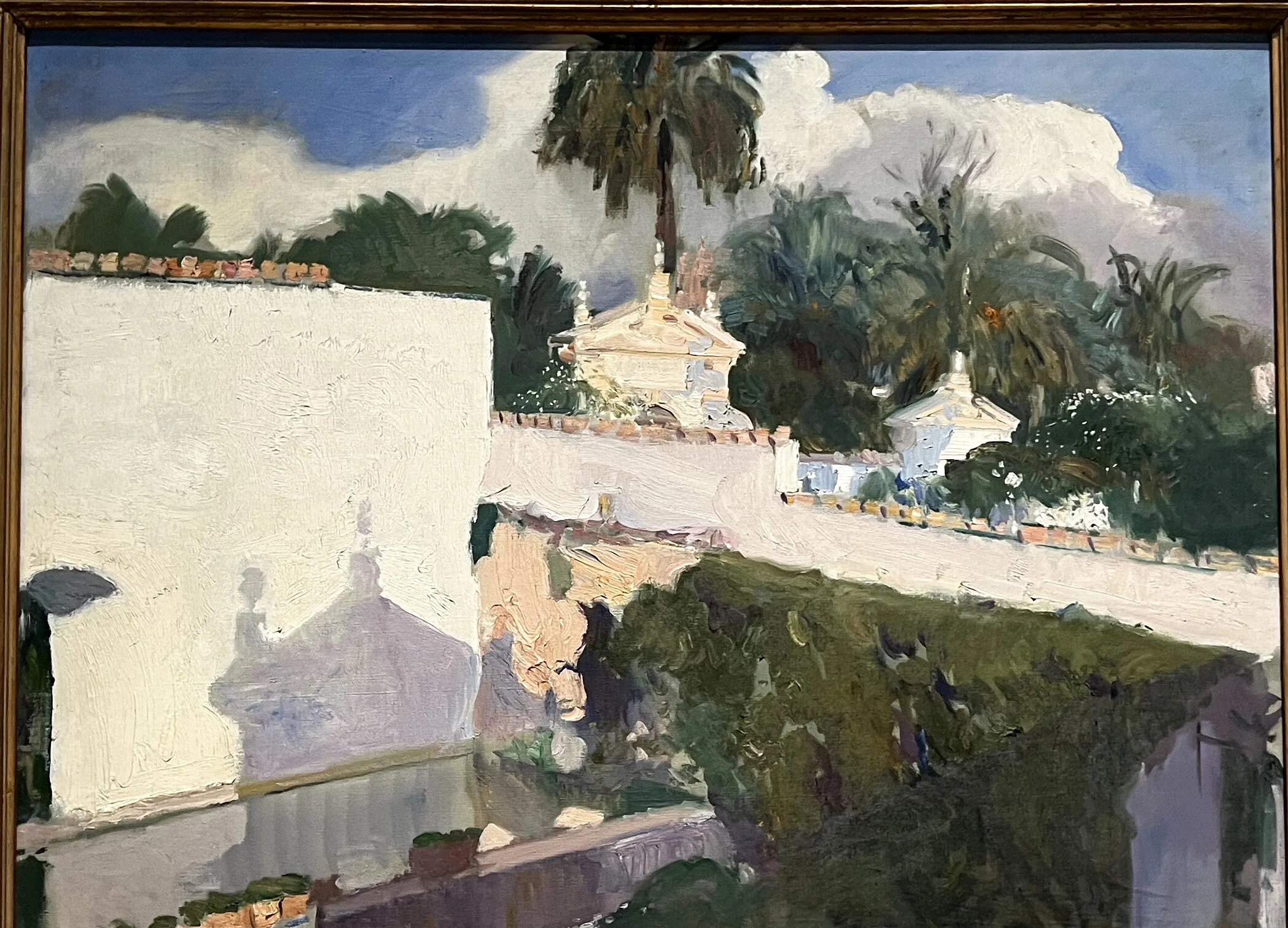 Sunlit whitewashed garden walls topped with terracotta tiles rise beside clipped hedges and a tall palm, their pale forms and cast shadows mirrored in a still Sevillian pool.