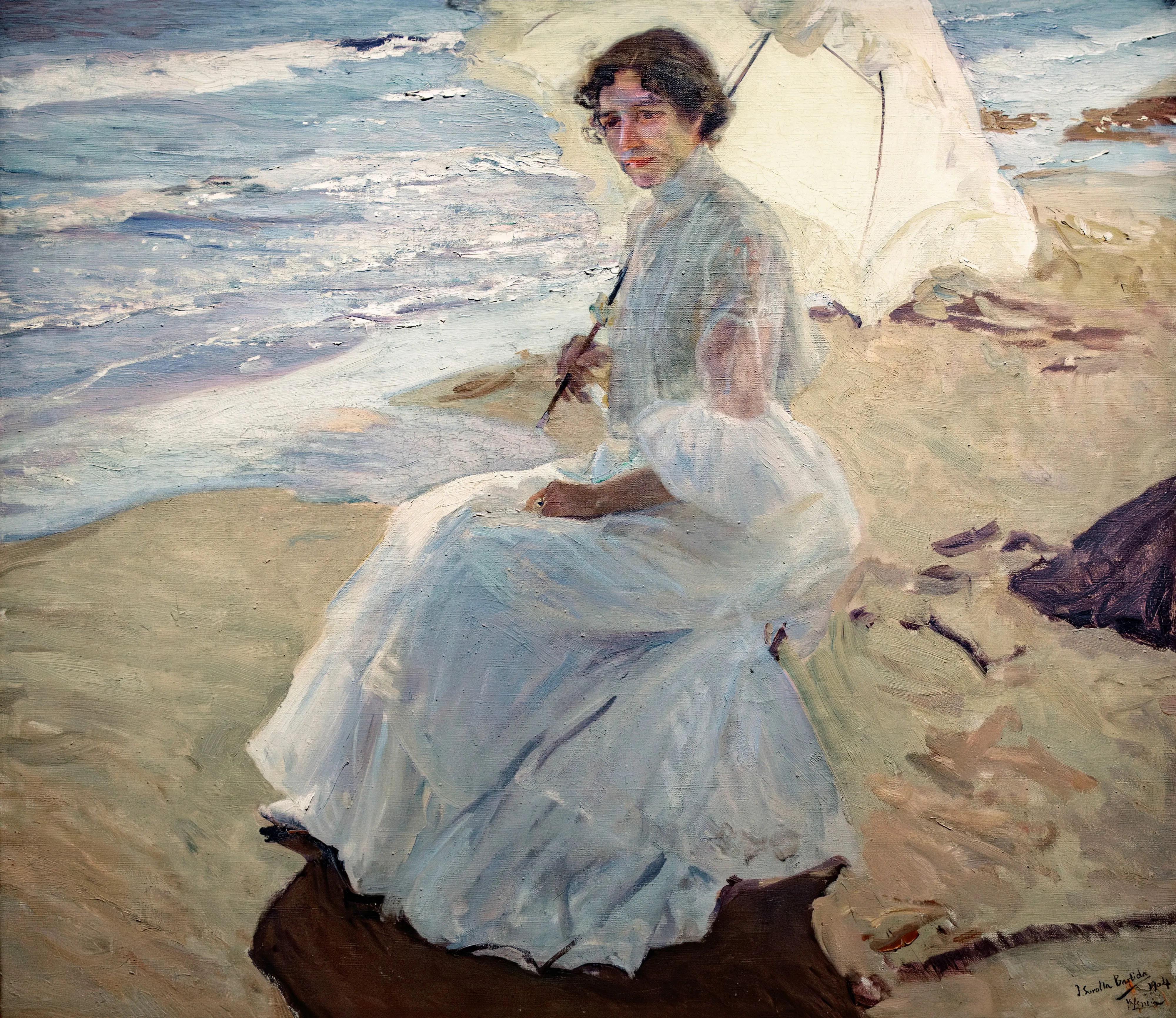 An elegant woman in a long white dress sits on the sunlit sand beneath a white parasol, the sea breaking behind her in clear coastal light.