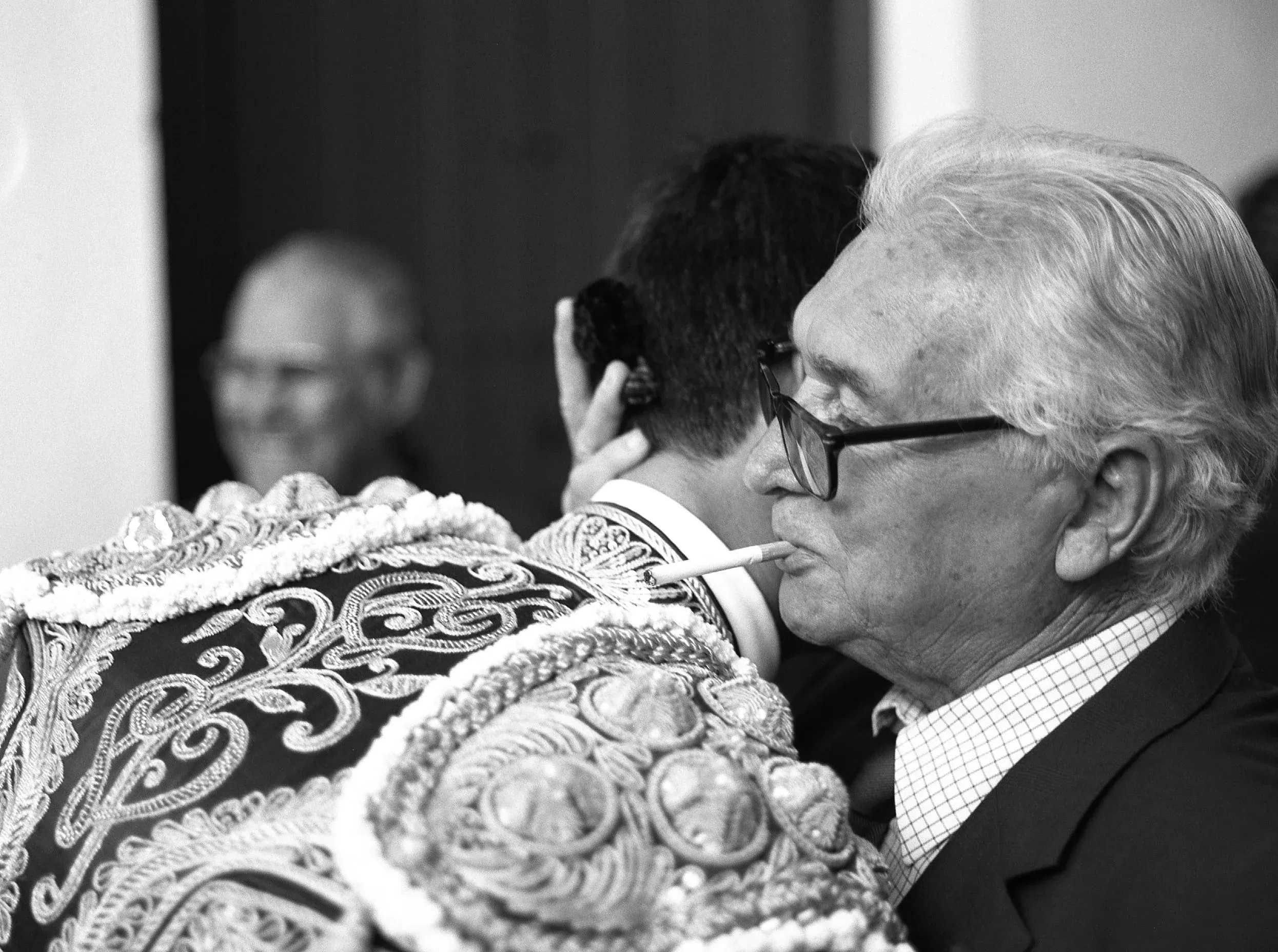 Close view of a matador’s embroidered suit of lights beside an older man with a cigarette.