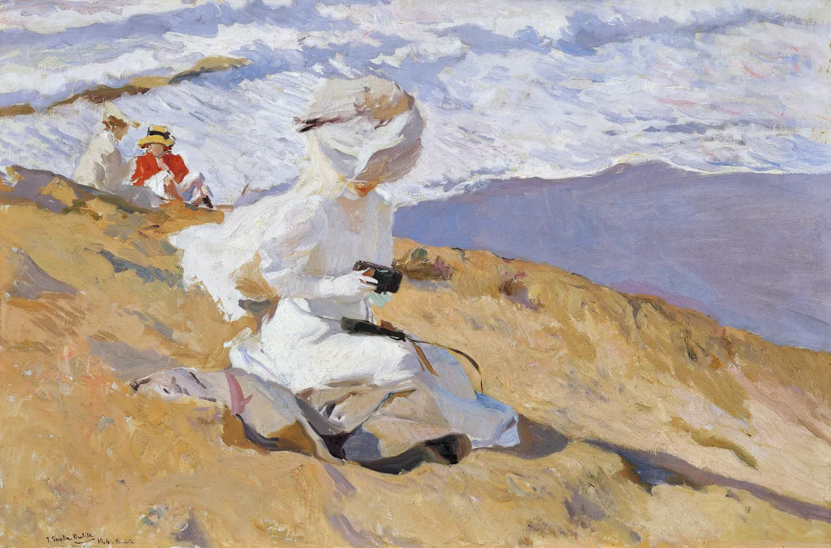 A woman in white with a wide summer hat sits on a windswept dune holding a small box camera, two children resting behind her above the glittering blue sea at Biarritz.