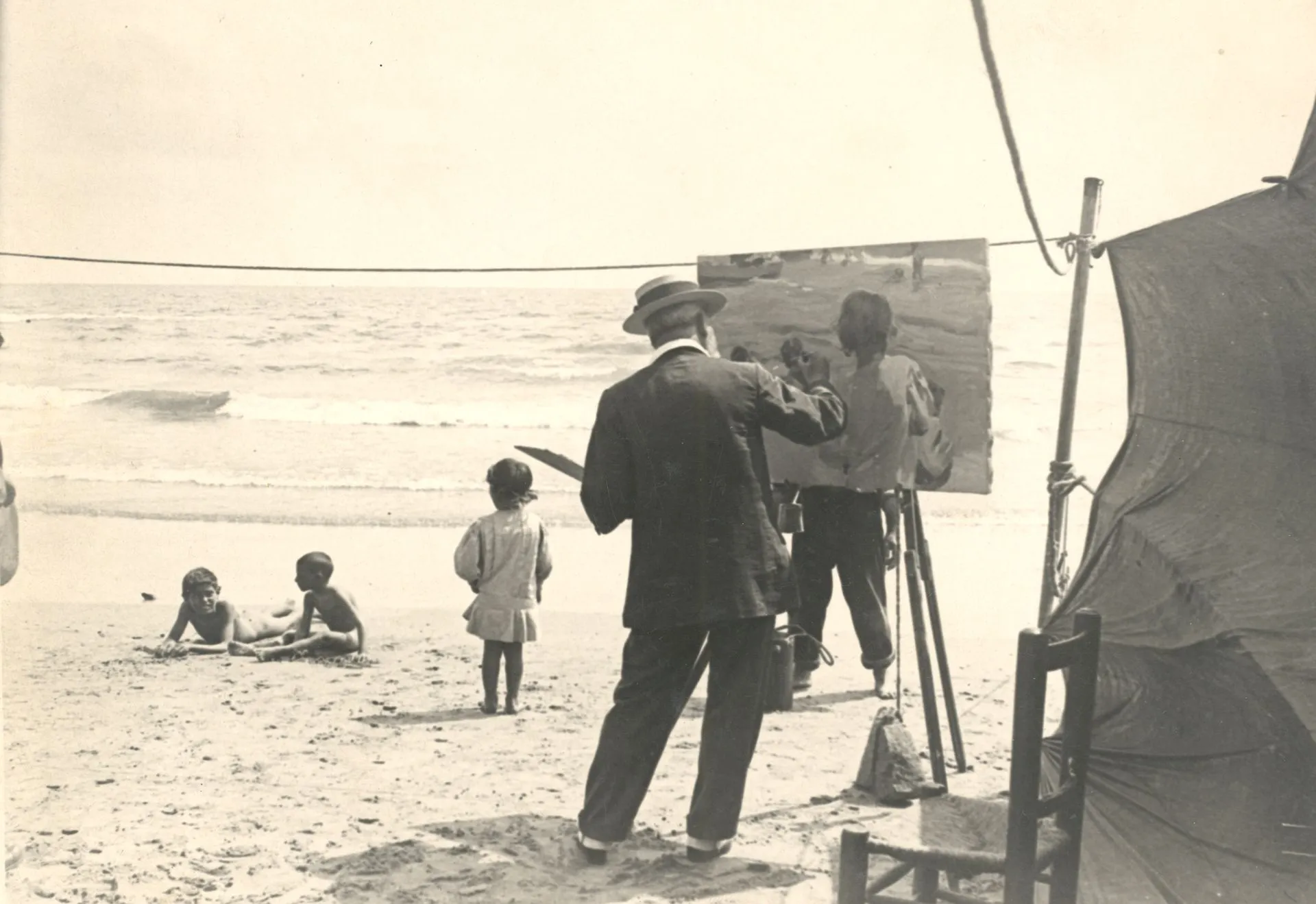 Black-and-white photograph of Joaquín Sorolla painting at his easel on the beach at El Cabañal, Valencia, in 1916, with two boys lying on the sand as his live models