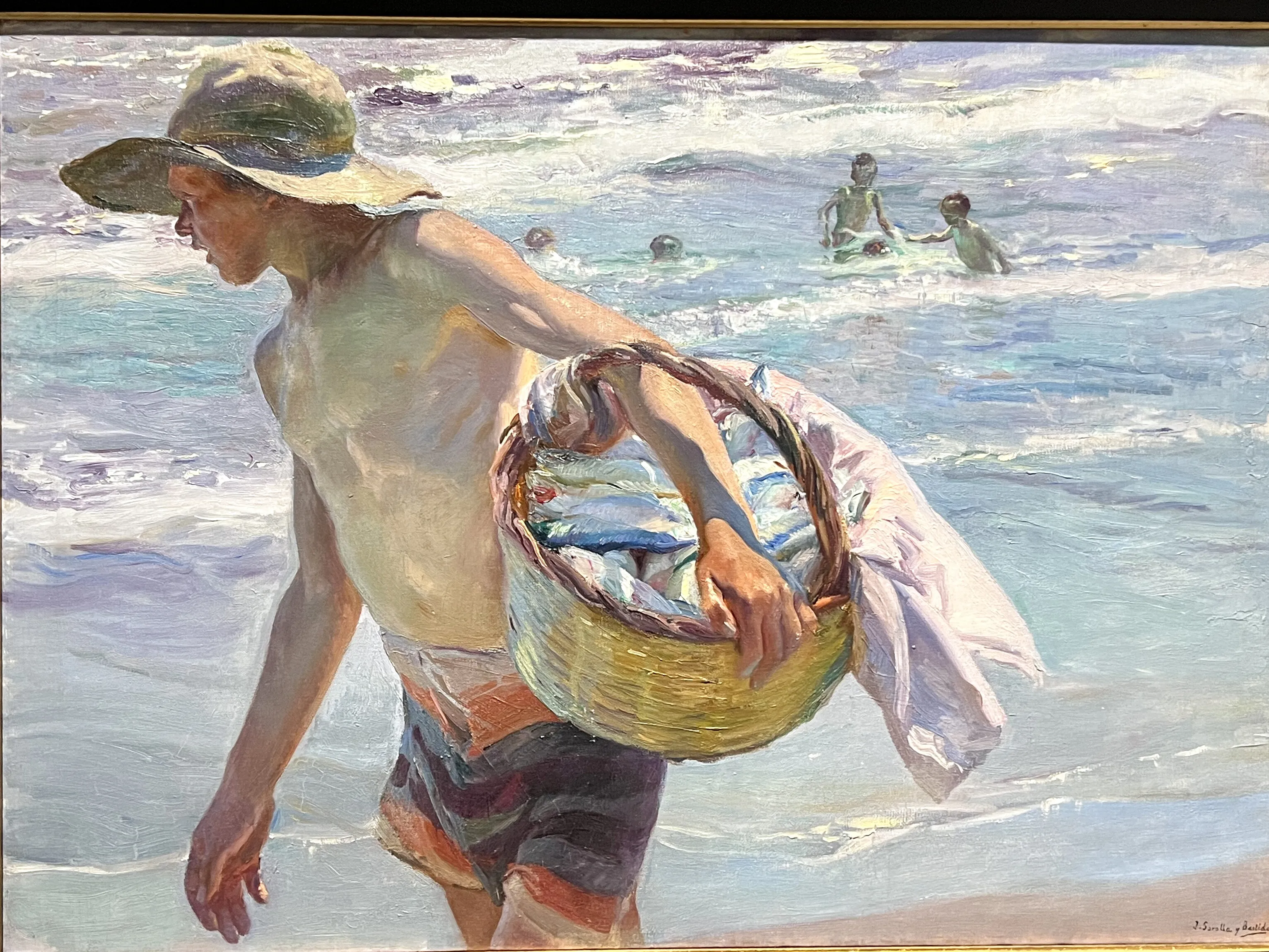 A bare-chested boy in a broad straw sun-hat strides across the wet Valencian sand carrying a wicker basket brimming with silvery fish, small bathers splashing in the sunlit surf behind him.